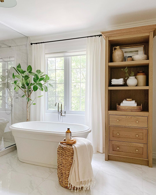 a glossy white acrylic bathtub in a white bathroom with a wooden cabinet & woven basket