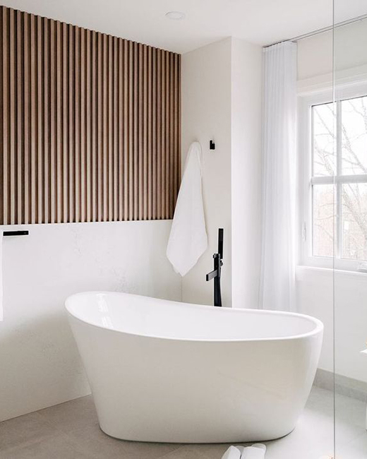 a glossy white acrylic bathtub in the corner of a white bathroom, with a dark wood paneled accent wall